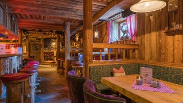 A cozy restaurant with wooden walls and rustic decor. There are table groups and a bar with stools in the foreground.