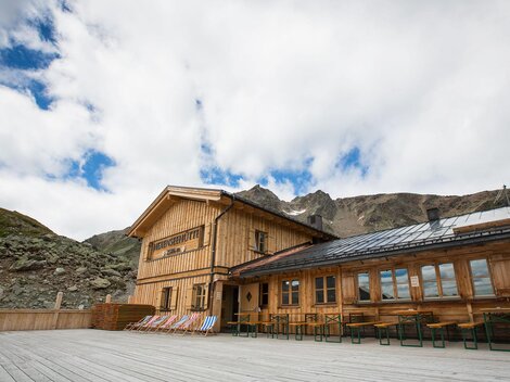 A rustic wooden house in the mountains with large windows. The sky is overcast and the surroundings are natural and green.