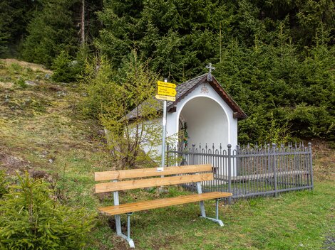 A small chapel stands in nature, surrounded by green trees. Next to the chapel is a wooden bench for sitting and resting. | © Bernhard Gruber – Photography
