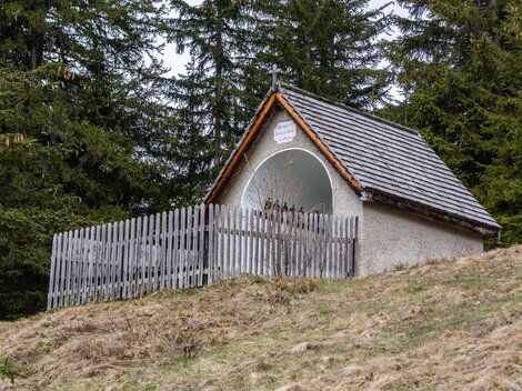 A small hut with a wooden fence in a wooded area. The sky is cloudy, and the ground is covered with grass and leaves. | © Bernhard Gruber – Photography