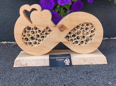 A decorative wooden sculpture in the shape of an infinity symbol with heart motifs and a spiral pattern. In the background, there are purple flowers.