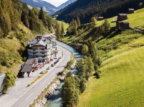 A picturesque landscape with a river flowing next to a road. In the background, green hills and some chalets can be seen.