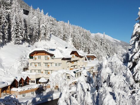 Ein malerisches Hotel in einer schneebedeckten Landschaft. Hohe Tannenbäume und blauer Himmel schaffen eine winterliche Atmosphäre.