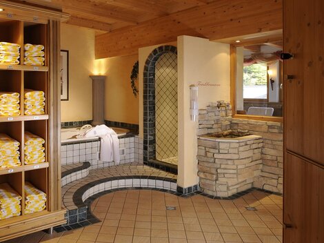 A relaxing wellness area with neatly stacked towels on shelves. In the background, a whirlpool and an inviting sauna can be seen.