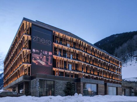A modern hotel building with wooden cladding and large windows. Surrounded by snow-covered mountains and a clear sky.