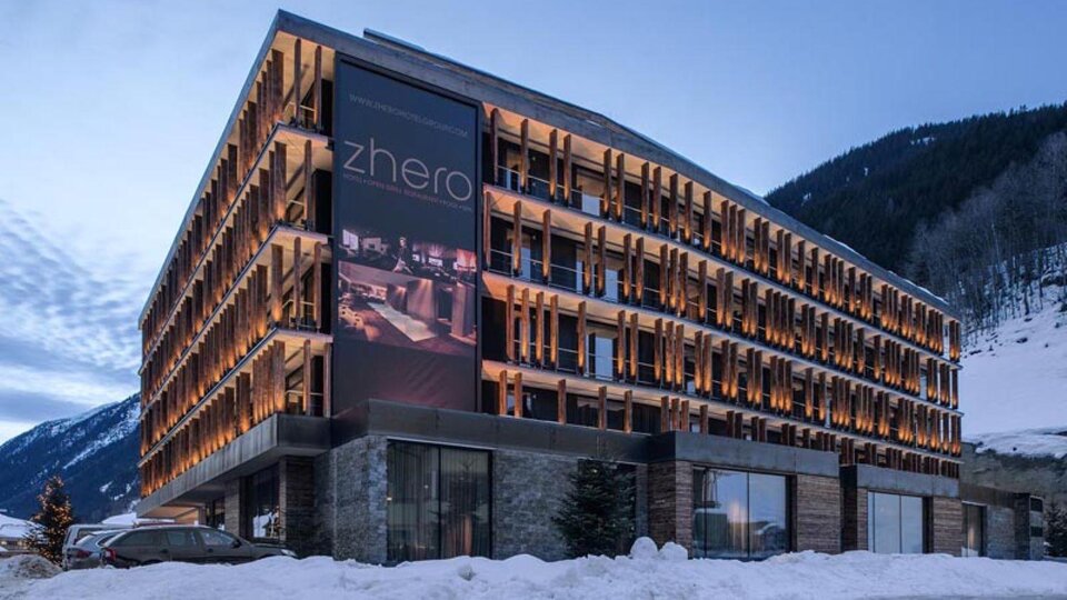 A modern hotel with a stylish facade and illuminated balconies. Surrounded by snow-covered mountains and a clear evening sky.