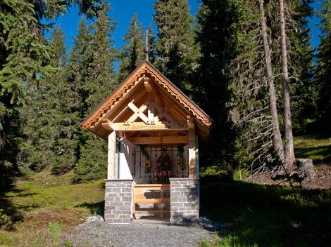 A small wooden chapel surrounded by tall trees. The sky is blue and the surroundings are green and peaceful. | © Bernhard Gruber – Photography