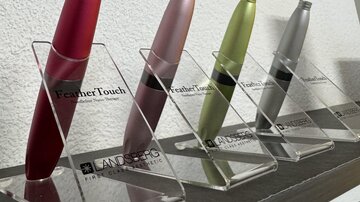 Four different cosmetic devices in various colors are displayed on a shelf. They are placed in transparent holders and showcase the brand "FeatherTouch".