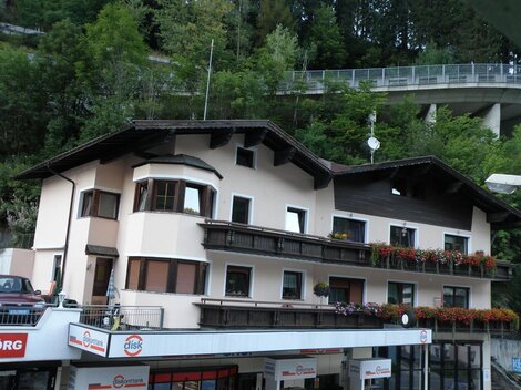 A charming building with several balconies and blooming windows. In the background, trees and a street bridge are visible.