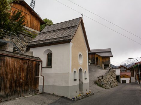 A small, white chapel stands on a narrow street, surrounded by traditional wooden houses. The sky is overcast and it seems to be a quiet, rural setting. | © Bernhard Gruber – Photography