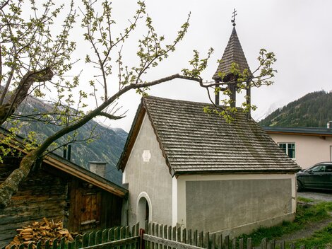 A small chapel with a pointed roof and a bell tower is surrounded by trees and mountains. In the foreground, there is a wooden fence. | © Bernhard Gruber – Photography