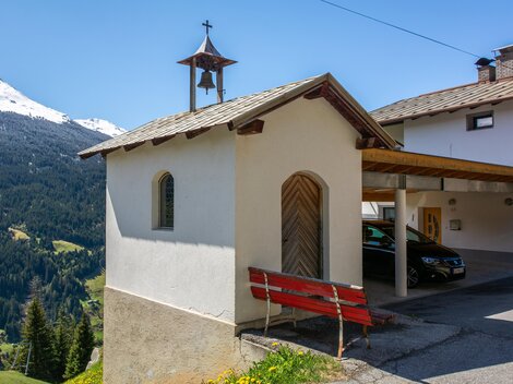 A traditional house with a small bell on the roof, surrounded by mountains. In the foreground, there is a red bench and some parked vehicles are visible. | © Bernhard Gruber – Photography