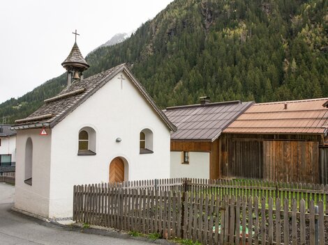 A small, white-painted church with a pointed roof stands in a rural setting. In the background, green mountains and traditional wooden houses can be seen. | © Bernhard Gruber – Photography