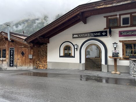 A traditional alpine building with an inviting entrance. Next to the building is a stack of wood and a sign that reads "STADL".
