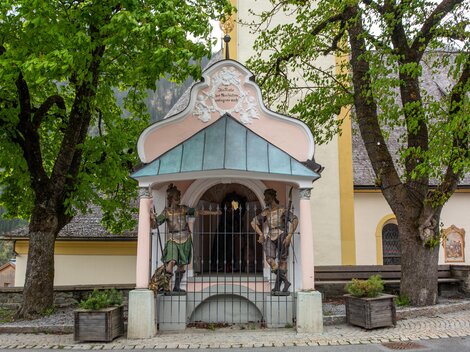 A small chapel with colorful figures and a green roof. In the background, there are large trees and a building with a yellow facade. | © Bernhard Gruber – Photography