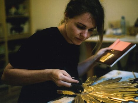 An artist is working on a gold-colored, star-shaped sculpture. She is holding a book in her hand and concentrating on her creative activity.