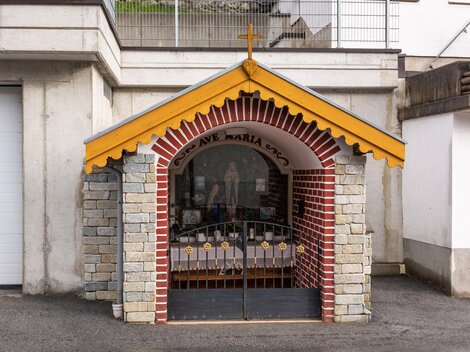 A small chapel with a yellow roof and a cross. It is built of stone and bricks and has a closed lattice door. | © Bernhard Gruber – Photography
