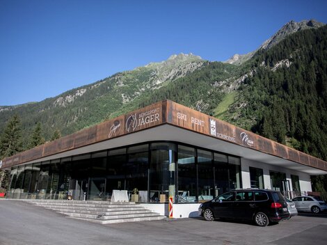 A modern building in the mountains with large windows. The surroundings are surrounded by green forests and mountains.
