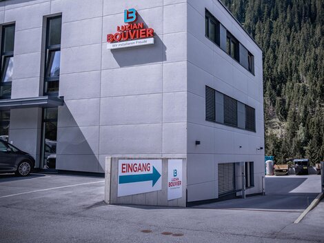 A modern building with a bright façade and the inscription "Bouvier". In the background, green mountains rise under a partly cloudy sky.