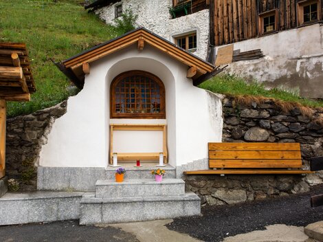 A small white building with a wooden window and a wooden bench.  
Surrounded by stones and green grass, it conveys a cozy, rural atmosphere.
