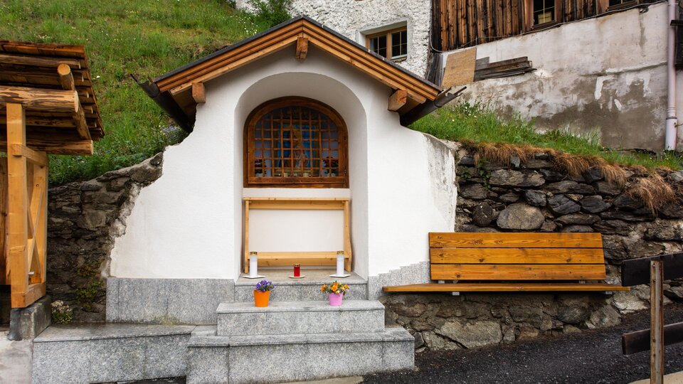 A small white building with a wooden window and a wooden bench.  
Surrounded by stones and green grass, it conveys a cozy, rural atmosphere.