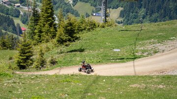 Ein Fahrzeug fährt eine kurvenreiche Schotterstraße im Gebirge. Im Hintergrund sind grüne Wiesen und Bäume sichtbar, während die Berglandschaft die Szenerie umgibt.