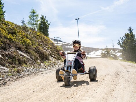 A child is riding a vehicle on a dirt road in the mountains. In the background, trees and a sunny landscape can be seen.