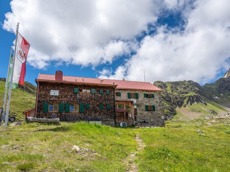 A stone mountain hut in the midst of a green meadow. In the background, mountains and a blue sky with some clouds can be seen.