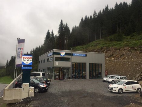 A modern service building by Bosch, surrounded by forests and mountains. Several cars are parked in the parking lot.
