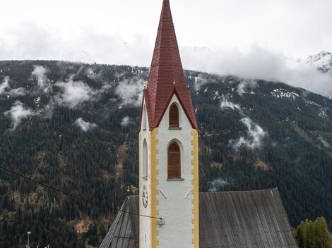 A church with a red roof and a tall bell tower in front of a mountainous landscape. The surroundings are characterized by green forests and gentle hills. | © Bernhard Gruber – Photography
