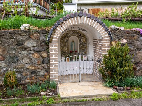 A small altar shrine in a stone wall with a white gate. Surrounded by green plants and flowers. | © Bernhard Gruber – Photography