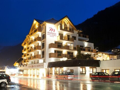 A modern hotel building with several stories and balconies. The surroundings are illuminated in the evening, and the streets are wet from the rain.