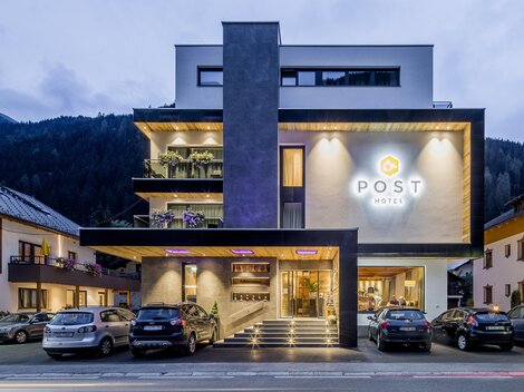 A modern hotel building with the inscription "POST" glows in the twilight. Surrounded by mountains and other buildings, it is an inviting place to stay.