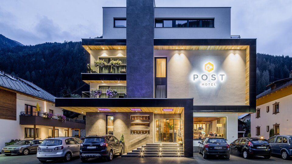 A modern hotel building with the inscription "POST" glows in the twilight. Surrounded by mountains and other buildings, it is an inviting place to stay.