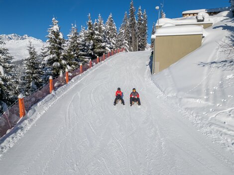 Two people are sitting on a snowy slope. Surrounded by snow-covered trees and a clear blue sky.