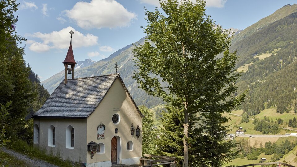 A small chapel surrounded by trees and mountains. The sky is partly cloudy and the landscape is green and picturesque.