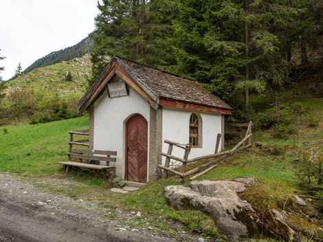 A small, picturesque cabin stands in the midst of a green landscape. Surrounded by trees and mountains, it offers a tranquil atmosphere. | © Bernhard Gruber – Photography