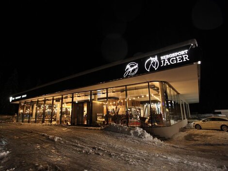 A modern sports shop with large windows, well-lit at night. It is surrounded by snow and the store name "Bergsport Jager" is clearly visible.