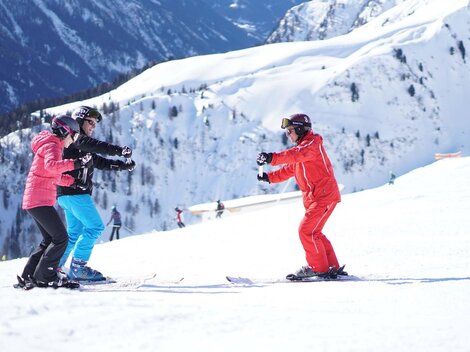 A ski school in the mountains with three people learning to ski. The surroundings are covered in snow and the mountains are impressive.