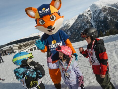 Ein Maskottchen in Form eines Fuchses steht auf einem Schneehang und interagiert mit drei Kindern. Die Kinder tragen Skiausrüstung und lachen.