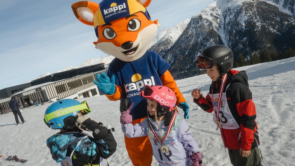 Ein Maskottchen in Form eines Fuchses steht auf einem Schneehang und interagiert mit drei Kindern. Die Kinder tragen Skiausrüstung und lachen.