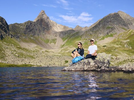 Two people are sitting on a rock at the edge of a lake in the mountains. The landscape is majestic with high peaks and a clear sky.