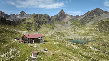 A picturesque mountain landscape with a cabin and a small lake. Surrounded by high mountains and green grass.