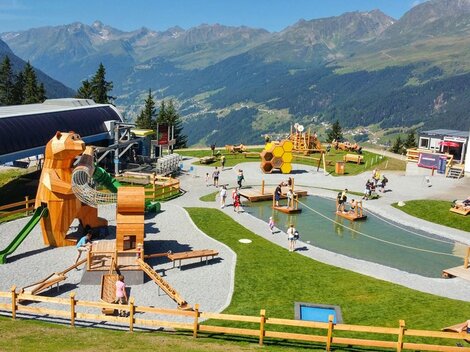 A playground in the mountains with various wooden play equipment for children. In the background, green meadows and tall mountains can be seen.