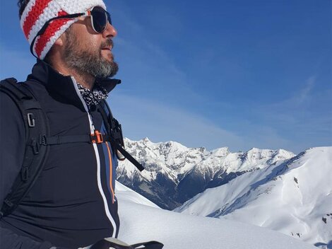 A man with sunglasses and a knit hat stands on a snow-covered mountain peak. In the background, majestic, snow-covered mountains can be seen under a clear blue sky.