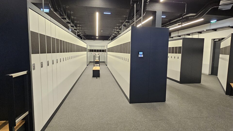 A modern locker room with many white lockers. The walls are gray and the floor is carpeted.