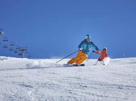 A skier is dynamically descending the slope while a child in orange ski clothing follows him. The sky is bright blue and the slope is covered with fresh snow.