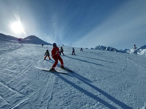 A group of skiing individuals on a sunny slope. Snow-covered mountains can be seen in the background.