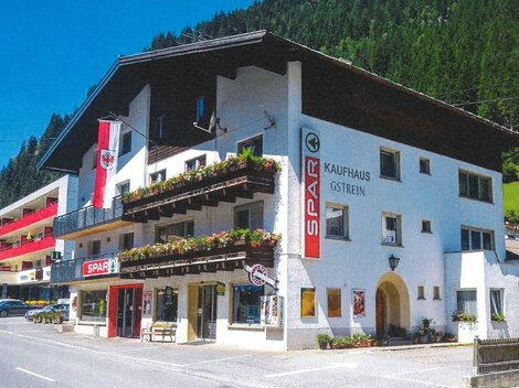 A small commercial building in alpine style with a discount supermarket on the ground floor. The facade is decorated with flower balconies and flags, surrounded by green mountains.