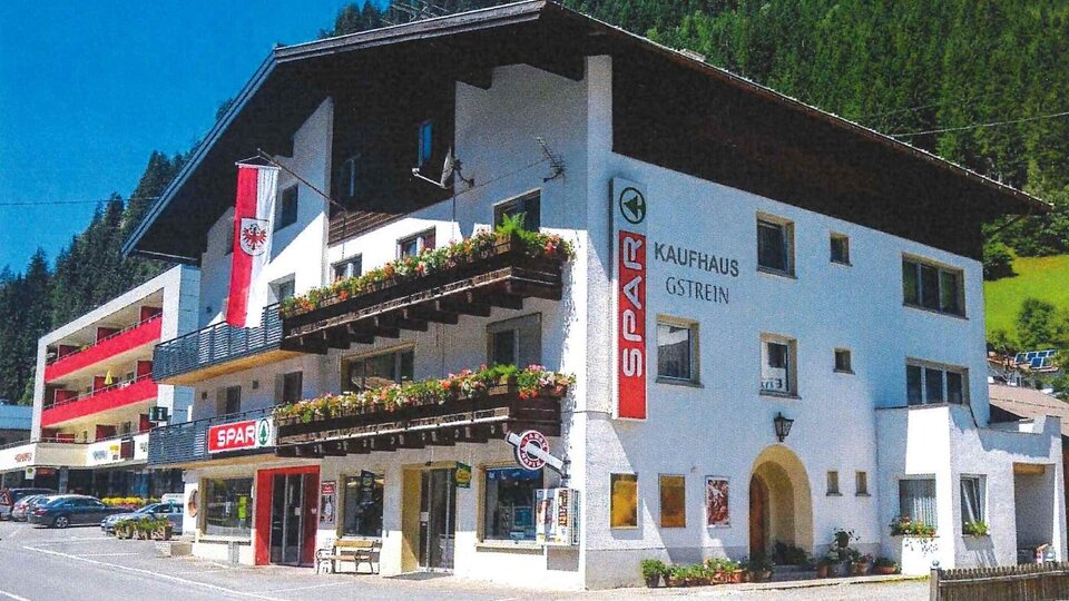 A small commercial building in alpine style with a discount supermarket on the ground floor. The facade is decorated with flower balconies and flags, surrounded by green mountains.